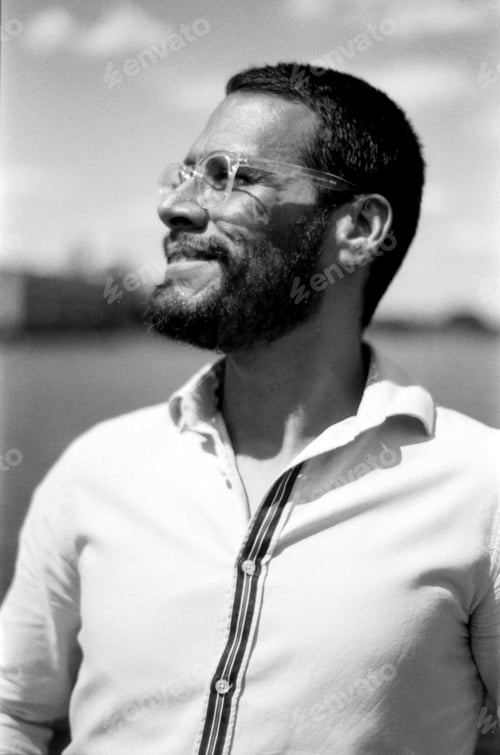 Preview: Bearded man wearing glasses smiling and looking up in monochrome analog photo