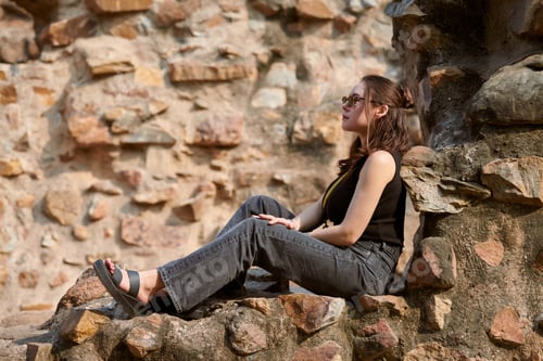 Preview: Attractive east asian woman in black sunglasses and black clothes sitting on ancient stone wall