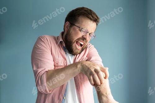 Preview: Unhappy annoyed man scratches itchy skin on arm and screams from the unpleasant sensation