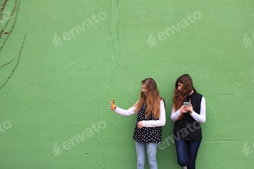 Preview: Two teens by monochrome green background wall snapping cell phone photos for social media sharing