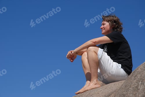 Preview: Woman outdoors in summer, sitting on a rock, thinking.