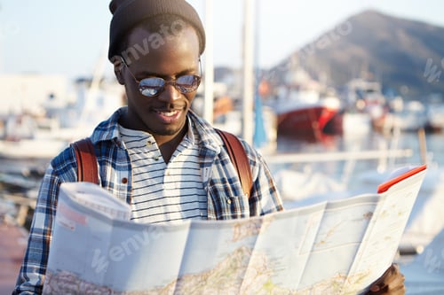 Preview: Outdoor shot of cheerful and stylish young dark-skinned European male traveler with knapsack studyin