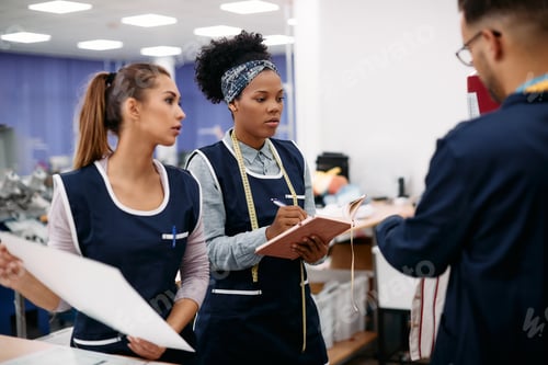Preview: Black tailor taking notes while working with her colleagues at textile factory.