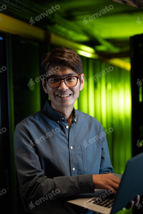 Preview: Portrait of asian male engineer smiling while using laptop in computer server room