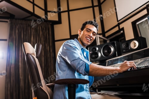 Preview: cheerful mixed race sound producer sitting in office chair in recording studio and looking at camera