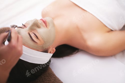 Preview: Beautiful young woman is getting facial clay mask at spa, lying with cucumbers on eyes