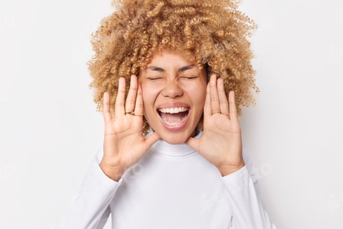 Preview: Overjoyed young curly woman keeps palms near face exclaims gladfully keeps eyes closed dressed in ca