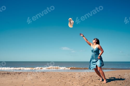 Preview: Young female in a summer dress trying to catch her hat in the air on the beach