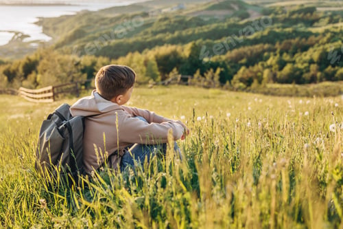 Preview: Rear view of tourist with backpack sitting on hill in grass field and enjoying beautiful landscape