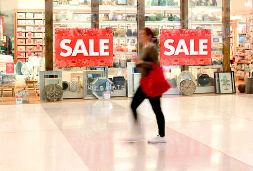 Preview: Woman walking past a retail store displaying sale signs in a shopping mall