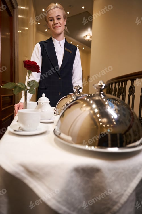 Preview: Smiling young woman on cart carrying hot food down hotel corridor
