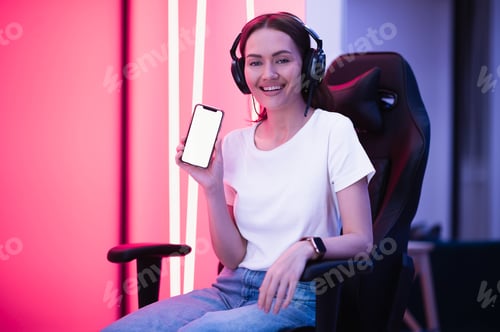 Preview: Happy promoter gamer girl wearing headset and sitting on gaming chair showing a blank smart phone