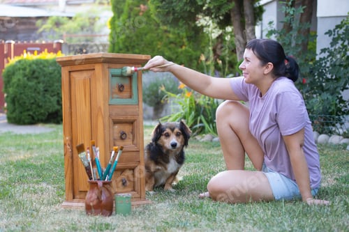 Preview: Woman painting a wooden furniture while sitting with her dog in the garden. DIY concept.