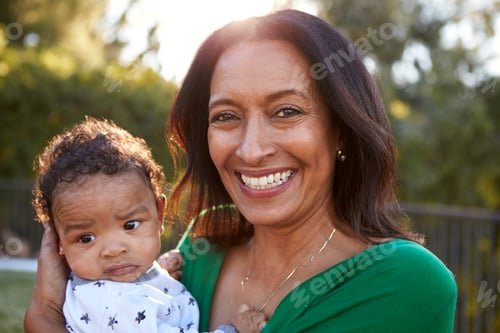 Preview: Happy mixed race middle aged grandmother holding her three month old grandson in garden