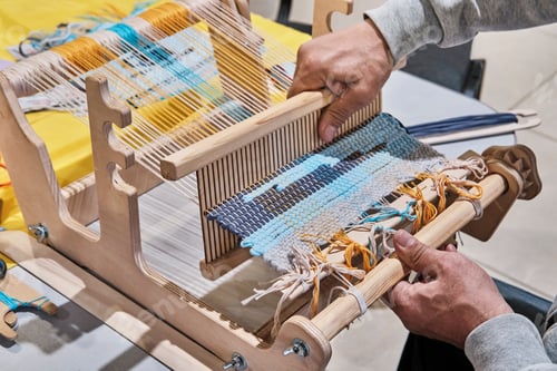 Preview: Hands of senior man weaving small rug with pattern on manual table loom, at masterclass on weaving.