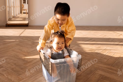 Preview: Cute Asian kids playing at home. A little boy carries his toddler sister in a plastic box.