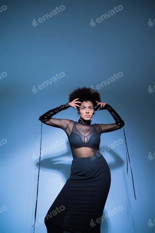 Preview: stylish african american woman in transparent shirt and black bra looking up on blue