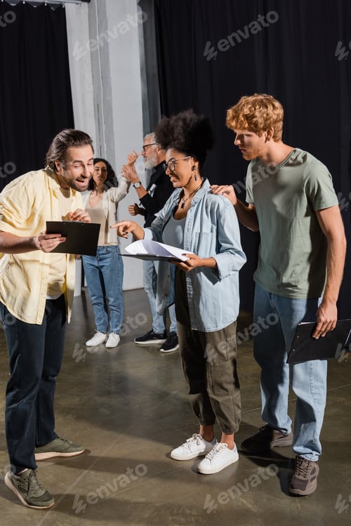 Preview: positive man showing clipboard with scenario to interracial actors on stage in theater
