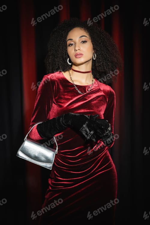 Preview: Stylish young african american woman in dress posing with handbag on burgundy drapery background