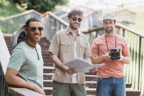 Preview: young man holding map and smiling at camera near carefree interracial friends on stairs on city
