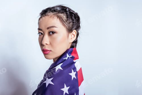 Preview: Portrait of stylish asian girl wrapped in american flag looking at camera isolated on grey