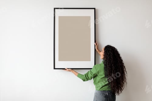Preview: Happy Woman Hanging Empty Poster In Frame On Wall Indoors