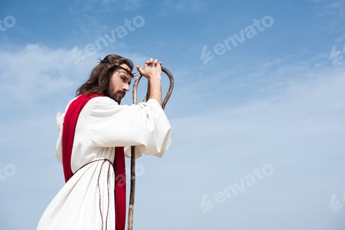 Preview: side view of Jesus in robe, red sash and crown of thorns leaning on wooden staff against blue sky