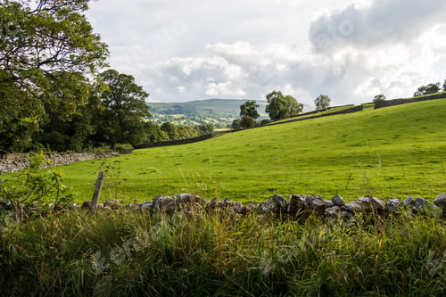 Preview: Pasture fenced with a stone wall against the background of forest and hills under a cloudy sky
