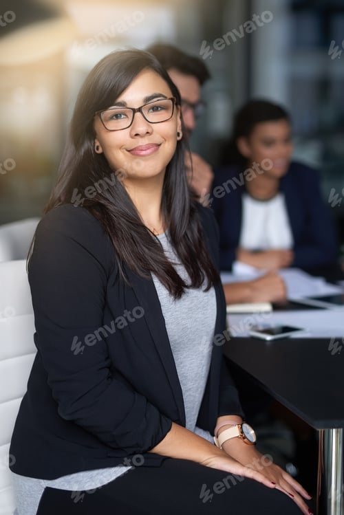 Preview: Portrait of a smiling young businesswoman sitting in an office with colleagues in the background