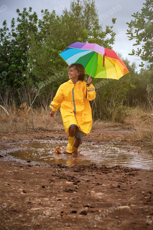 Preview: Smiling boy with rainbow umbrella playing in puddle