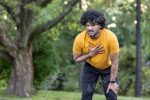 Preview: Tired young Indian male athlete standing bent over in park and holding hand to chest, feeling