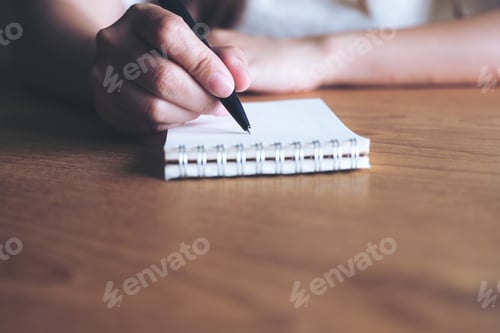 Preview: Closeup image of a woman's hand writing down on a white blank notebook on wooden table