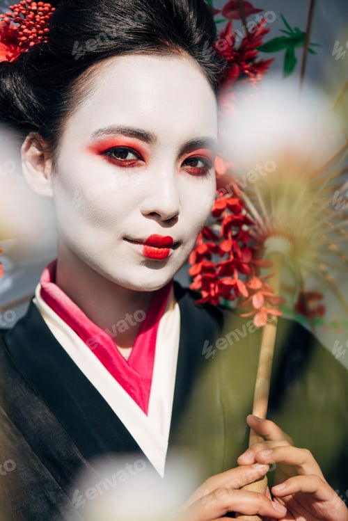 Preview: selective focus of tree branches and beautiful smiling geisha with umbrella in sunlight