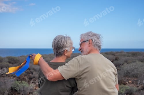 Preview: Rear view of senior couple looking into the eyes while man holds the colors of the Ukrainian flag