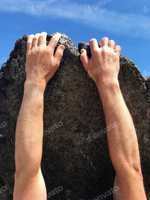 Preview: A pair of male hands and arms clinging by fingertips to side of a harsh rock cliff whilst climbing