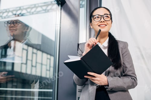 Preview: smiling businesswoman standing by window and holding notebook