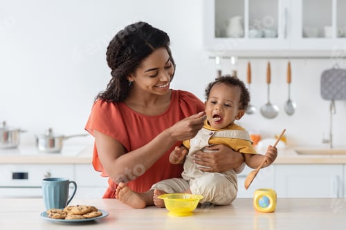 Preview: Childcare. Loving Black Mom Feeding Her Baby Son From Spoon In Kitchen