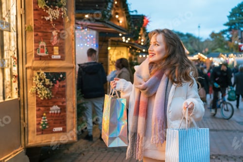 Preview: Happy woman enjoys shopping at festive Christmas market while carrying bags, walking in Xmas fair