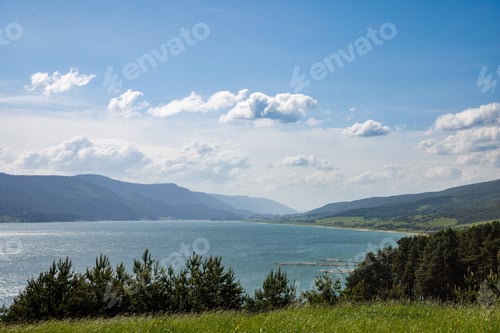Preview: A lake reflects sunlight, against blue sky and mountain ranges and villages
