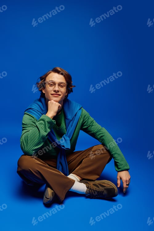Preview: Fashionable young man radiates style while sitting confidently against a vibrant blue backdrop