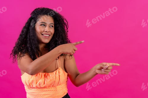 Preview: Curly-haired woman in summer clothes on a pink background pointing to the right, studio shot