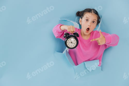 Preview: Surprised young girl pointing at alarm clock held in hand, emerging from torn blue paper background