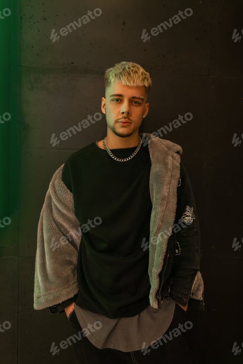 Preview: Trendy cool guy model with hair in a trendy black jacket stands and poses near a black background