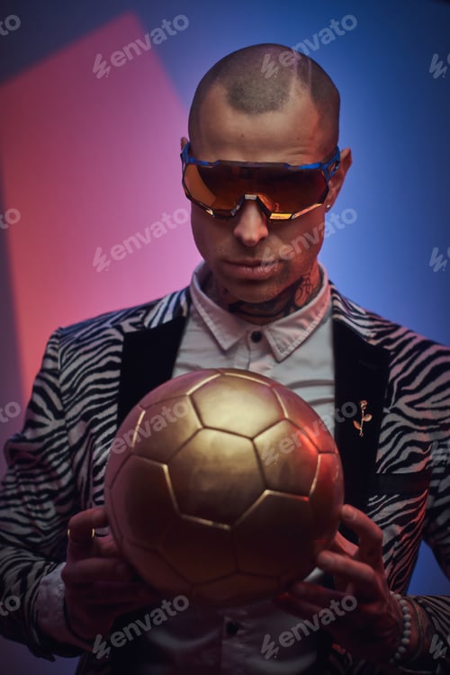 Preview: Elegant tattooed male model posing with a golden soccer ball in a studio