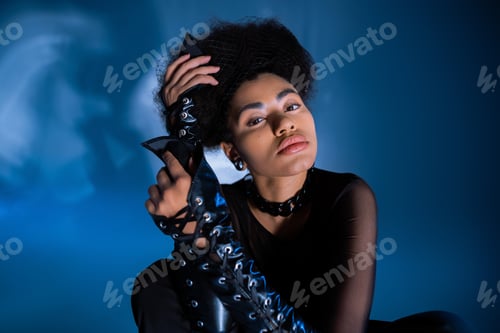 Preview: stylish african american woman in transparent shirt and lace-up leather sleeves posing on blue