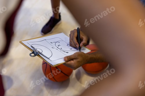 Preview: Biracial female basketball players with male coach discussing game plan on clipboard at gym