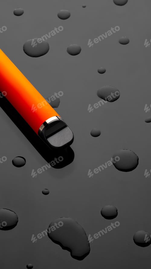 Preview: Close-up of an orange pen on a black surface with water droplets at midday