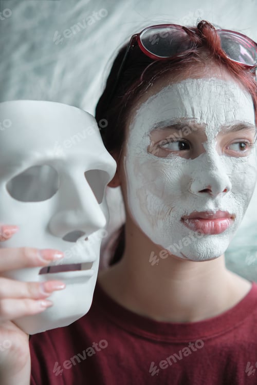 Preview: Teenage girl with a clay cosmetic mask applied to her face holds a white theater mask in hand.