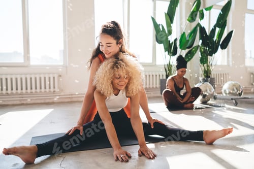 Preview: Flexible woman practicing yoga in gym with ethnic friend