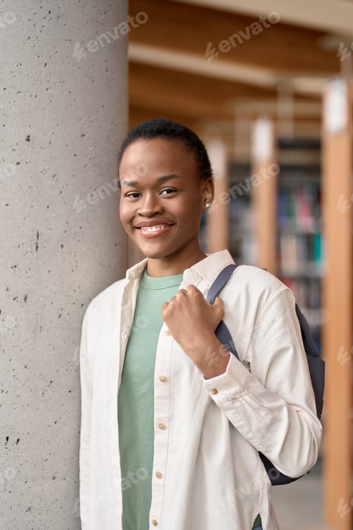 Preview: Happy African girl student holding backpack in library, vertical portrait.